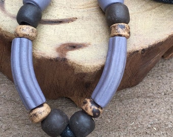 May include: A necklace with a brown and white ceramic pendant featuring a stylized figure. The necklace is made with purple, brown, and gold beads and a black cord.