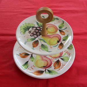 May include: Two-tiered white ceramic serving stand with a wooden handle. The stand is decorated with a hand-painted design of pears, grapes, and leaves.