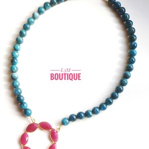 以下が含まれることがあります： 青と緑の宝石のネックレスに、ピンクの宝石のペンダントが付いている。ペンダントは、金色の枠で囲まれた円形をしている。ネックレスは丸いビーズでできており、金色の留め具が付いている。画像には「L3M BOUTIQUE」というテキストが表示されている。