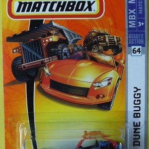 Puede incluir: Un coche de juguete Dune Buggy azul Matchbox con una jaula antivuelco roja. El coche está sobre un fondo amarillo y naranja con el logotipo de Matchbox y las palabras "MBX METAL" y "READY FOR ACTION".