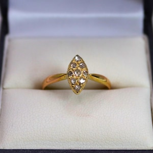 Puede incluir: Un anillo de oro con un grupo de pequeñas piedras preciosas transparentes en forma de marquesa. El anillo está engastado en una banda de oro y se presenta dentro de una caja de joyería negra con interior blanco.