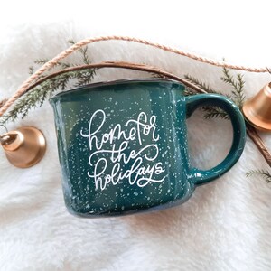 May include: A teal enamel mug with white speckles and the words "home for the holidays" in white script. A gold bell and sprigs of greenery are arranged around the mug on a white, fluffy surface.