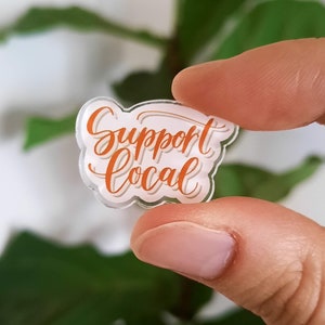 May include: A clear acrylic sticker with the text "Support Local" in orange lettering.