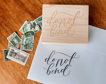 May include: A wooden stamp with the words "do not bend" carved into it. The stamp is sitting on a white envelope with the same words printed in black ink.