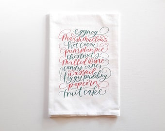 May include: White kitchen towel with a list of Christmas treats written in green and red calligraphy. The text reads: "eggnog, marshmallows, hot cocoa, pumpkin pie, chestnuts, mulled wine, candy canes, wassail, figgy pudding, popcorn, fruitcake"