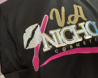 以下が含まれることがあります： ピンクとゴールドのロゴが施された黒いサロンケープ。ロゴには「VA NICHOL COSMETICS」とあり、リップスティックとメイクブラシのデザインが施されています。