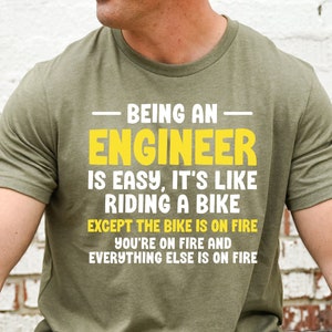 May include: A green t-shirt with a white graphic print that reads "Being an Engineer is easy, it's like riding a bike except the bike is on fire, you're on fire and everything else is on fire."