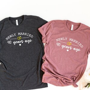 May include: Two heather grey t-shirts with white text that reads "Newly Married 40 years ago" and a graphic of two gold rings. The shirts are laid out on a white background.