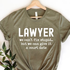 May include: Olive green t-shirt with the word "LAWYER" in large white letters. Below, the text reads "we can't fix stupid... but we can give it a court date." The shirt is displayed on a wooden hanger.
