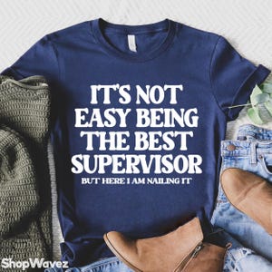 May include: A navy blue t-shirt with the white text: "IT'S NOT EASY BEING THE BEST SUPERVISOR BUT HERE I AM NAILING IT". The shirt is shown with jeans, a green sweater, and brown boots.