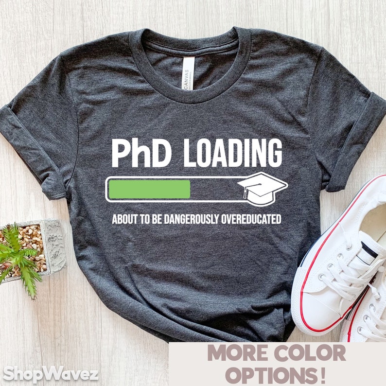 May include: Dark grey t-shirt featuring the text "PhD LOADING" above a progress bar graphic. The bar is partially filled with green, and a graduation cap icon is at the end. The phrase "ABOUT TO BE DANGEROUSLY OVEREDUCATED" is below the graphic.