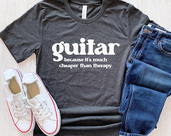 May include: A dark gray t-shirt with white text that reads "guitar because it's much cheaper than therapy". The shirt is laid flat on a light wood surface with a pair of white canvas shoes and blue jeans.
