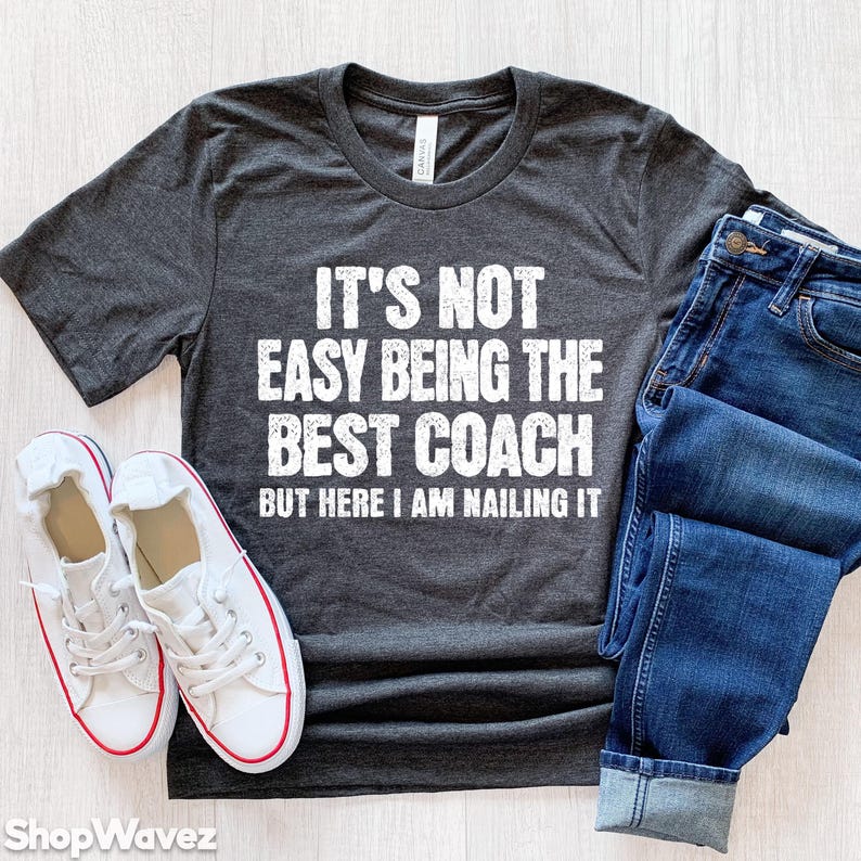 May include: A dark gray t-shirt with a white graphic that reads "It's not easy being the best coach but here I am nailing it".