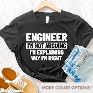 May include: Black t-shirt with white text that reads "ENGINEER I'M NOT ARGUING I'M EXPLAINING WHY I'M RIGHT". The t-shirt is folded and laid on a wooden surface with a pair of yellow sneakers and blue jeans.