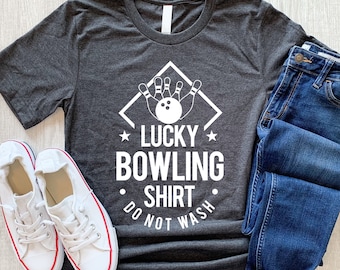 May include: A dark grey t-shirt with a white graphic of bowling pins and a bowling ball in a diamond shape. The text "Lucky Bowling Shirt Do Not Wash" is printed below the graphic.