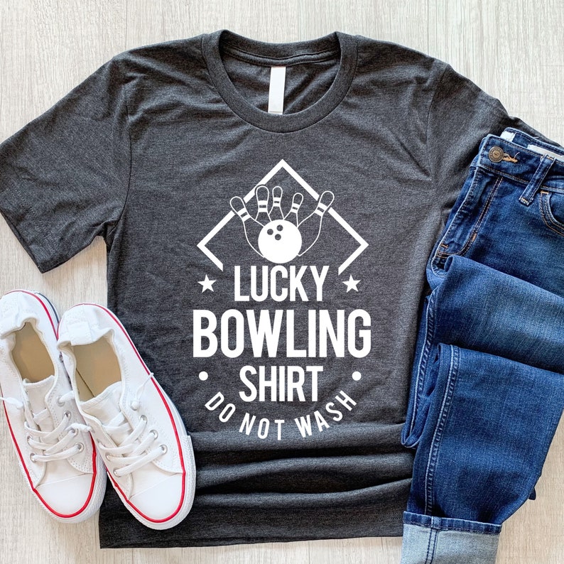 May include: A dark gray t-shirt with a white graphic of bowling pins and a bowling ball in a diamond shape. The text "Lucky Bowling Shirt Do Not Wash" is printed below the graphic.