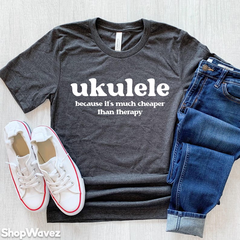 May include: A dark gray t-shirt with the text "ukulele because it's much cheaper than therapy" in white. The shirt is laid flat on a light wood surface with a pair of white sneakers and blue jeans.
