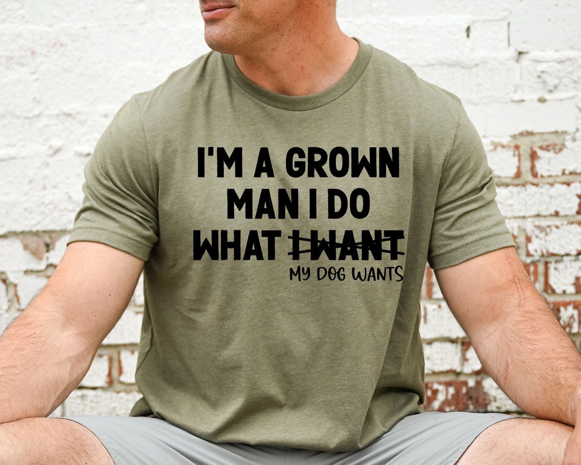 A Dog Dad wearing an olive green graphic t-shirt that says "I'm A Grown Man I Do What My Dog Wants".
