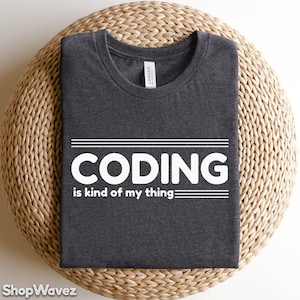 May include: Dark gray t-shirt with the text "CODING is kind of my thing" in white. The shirt is folded and placed on a woven, circular surface. The shirt has a crew neck and is made of a soft material.