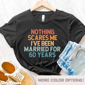 May include: A black t-shirt with a vintage-style graphic that reads "Nothing scares me I've been married for 60 years". The graphic is in shades of orange, yellow, blue, and green. The t-shirt is displayed on a wooden surface with a pair of yellow sneakers and a pair of blue jeans.