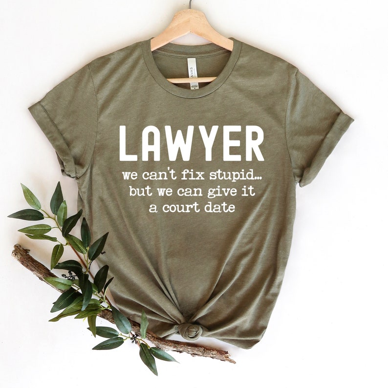 May include: Olive green t-shirt with the word "LAWYER" in large white letters. Below, the text reads "we can't fix stupid... but we can give it a court date." The shirt is displayed on a wooden hanger.