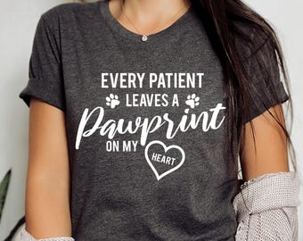 May include: A grey t-shirt with the text "Every Patient Leaves a Pawprint on My Heart" in white lettering. The text is surrounded by two paw prints. A heart with the word "Heart" inside is below the text.