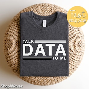 May include: Dark grey t-shirt with the white text "TALK DATA TO ME" across the front. The shirt is displayed on a woven, light brown surface. A yellow circle with "Fast shipping" is in the upper right corner. The t-shirt is made of a soft material.