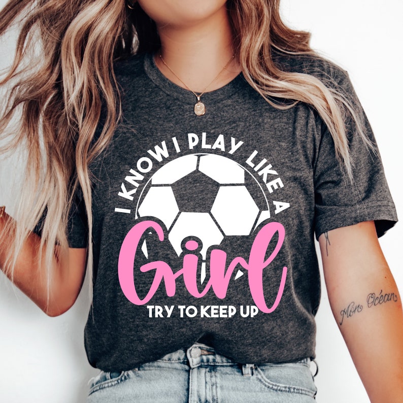 Puede incluir: Una camiseta gris con un bal&oacute;n de f&uacute;tbol blanco y el texto "I know I play like a girl try to keep up" en blanco con la palabra "girl" en rosa.