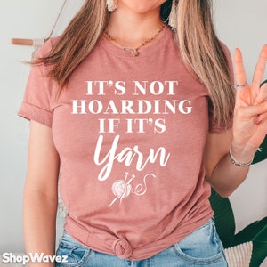 Op de afbeelding: Een vrouw draagt een roze T-shirt met de tekst "Het is geen hamsteren als het garen is" en een afbeelding van garen en breinaalden.