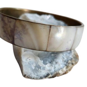 May include: A white and gold bangle bracelet with a mother of pearl inlay. The bracelet is sitting on a white and gray rock.