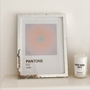 May include: A silver framed print with a gradient of pink, orange, and blue colors. The print has the text "PANTONE 523 AURA" at the bottom. A white candle with the text "NEW YORK CITY" and the brand name "homesick" is next to the print.