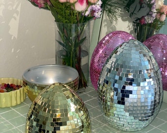 May include: Three disco ball shaped ornaments, one gold, one blue, and one pink, on a light blue tiled surface. A bouquet of pink, white, and yellow flowers in a glass vase is in the background.