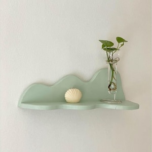 May include: A light green, wavy shelf with a white seashell and a glass vase with a small plant. The shelf is mounted on a white wall.