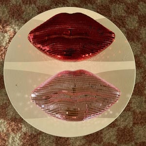 May include: Two decorative lip sculptures, one red and one pink, each covered in small, reflective tiles. The lips are mounted on a white, circular base, creating a striking visual contrast. The background features a patterned rug.