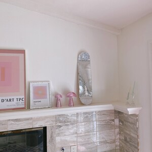 Puede incluir: Una repisa de chimenea de azulejos blancos con un espejo plateado, dos l&aacute;mparas de hongo rosa y una impresi&oacute;n enmarcada con la palabra "Pantone" en ella. La repisa est&aacute; sobre una chimenea negra.