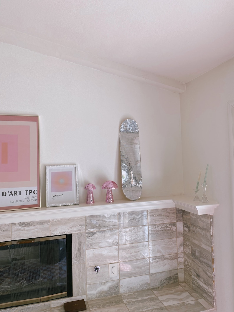 Puede incluir: Una repisa de chimenea de azulejos blancos con un espejo plateado, dos l&aacute;mparas de hongo rosa y una impresi&oacute;n enmarcada con la palabra "Pantone" en ella. La repisa est&aacute; sobre una chimenea negra.