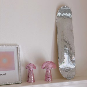 Puede incluir: Dos l&aacute;mparas de champi&ntilde;&oacute;n de mosaico rosa y una tabla de skate de mosaico plateado en un estante blanco con una pared blanca en el fondo.