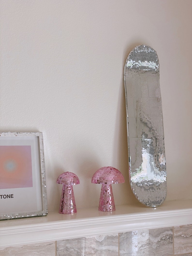Puede incluir: Dos l&aacute;mparas de champi&ntilde;&oacute;n de mosaico rosa y una tabla de skate de mosaico plateado en un estante blanco con una pared blanca en el fondo.