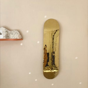 Puede incluir: Un espejo dorado con forma de tabla de skate. El espejo est&aacute; hecho de peque&ntilde;as baldosas cuadradas que crean un efecto de mosaico.