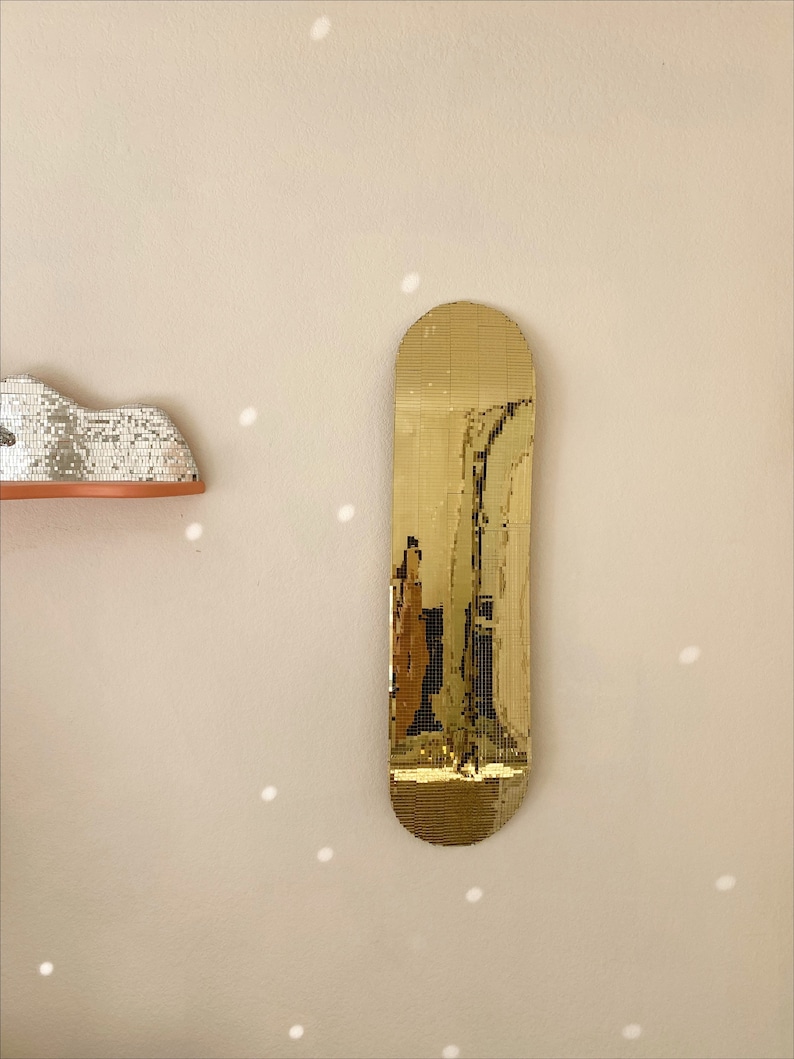 Puede incluir: Un espejo dorado con forma de tabla de skate. El espejo est&aacute; hecho de peque&ntilde;as baldosas cuadradas que crean un efecto de mosaico.