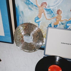 Peut inclure: Un tourne-disque vintage avec un disque vinyle noir sur le plateau tournant. Le tourne-disque est bleu et porte l'inscription "CROSLEY" sur le devant. Il y a un disque argenté de style mosaïque sur la table à côté du tourne-disque.
