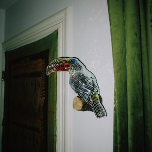 May include: A decorative toucan sculpture made of reflective, mirrored pieces. The bird is perched on a wooden branch against a white wall, with a colorful beak and a tail of black and silver. Green curtains and a wooden door are in the background.
