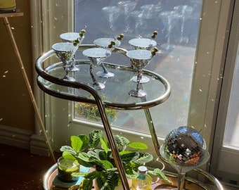 May include: A gold bar cart with two tiers. The top tier has six silver martini glasses with green olives. The bottom tier has a green plant, a candle, a bottle of liquor, and a silver disco ball.