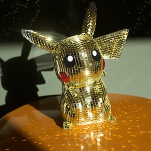 May include: A golden Pikachu figurine covered in small, reflective squares, creating a disco ball effect. The figure has black eyes, red cheeks, and pointed ears. It sits on a glossy, orange surface.