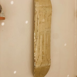 Puede incluir: Una tabla de skate dorada con una superficie de espejo. La tabla de skate est&aacute; colgada en una pared.