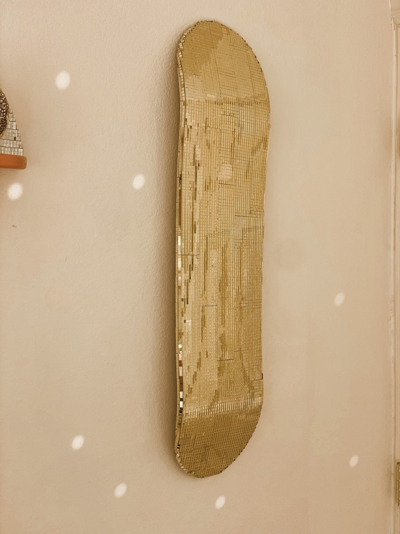 Puede incluir: Una tabla de skate dorada con una superficie de espejo. La tabla de skate est&aacute; colgada en una pared.