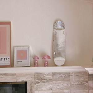 Puede incluir: Una repisa de chimenea de azulejos blancos con una patineta de espejo plateado, dos l&aacute;mparas rosadas en forma de hongo y dos impresiones enmarcadas. Una impresi&oacute;n es una muestra de color Pantone y la otra es un dise&ntilde;o abstracto rosa y blanco.