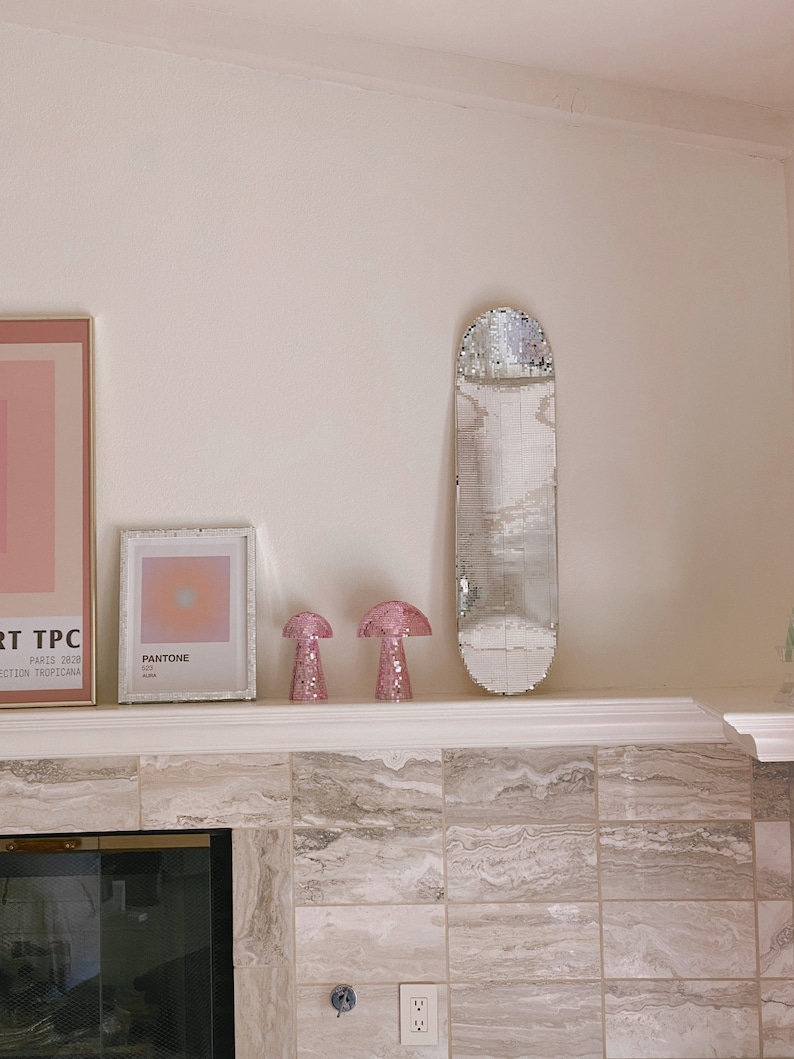 Puede incluir: Una repisa de chimenea de azulejos blancos con una patineta de espejo plateado, dos l&aacute;mparas rosadas en forma de hongo y dos impresiones enmarcadas. Una impresi&oacute;n es una muestra de color Pantone y la otra es un dise&ntilde;o abstracto rosa y blanco.