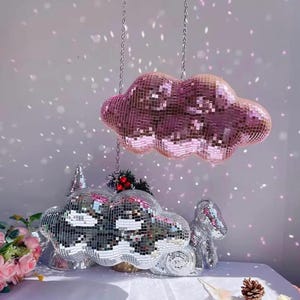 Könnte beinhalten: Eine rosa Discokugel-Wolke hängt an einer Kette über einer Sammlung von wolkenförmigen Discokugel-Skulpturen. Die Skulpturen sind silberfarben und stehen auf einer weißen Oberfläche.