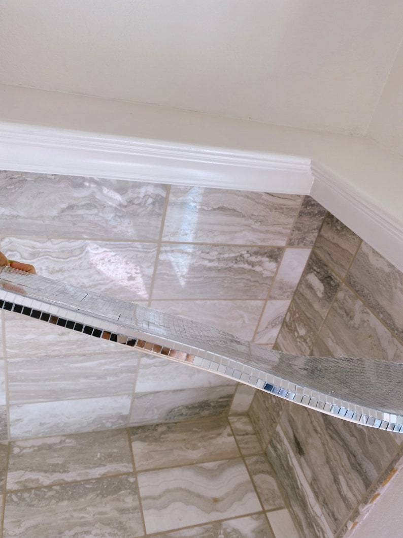 Puede incluir: Un borde de azulejos de espejo plateado se est&aacute; instalando en un ba&ntilde;o con azulejos de m&aacute;rmol gris claro.